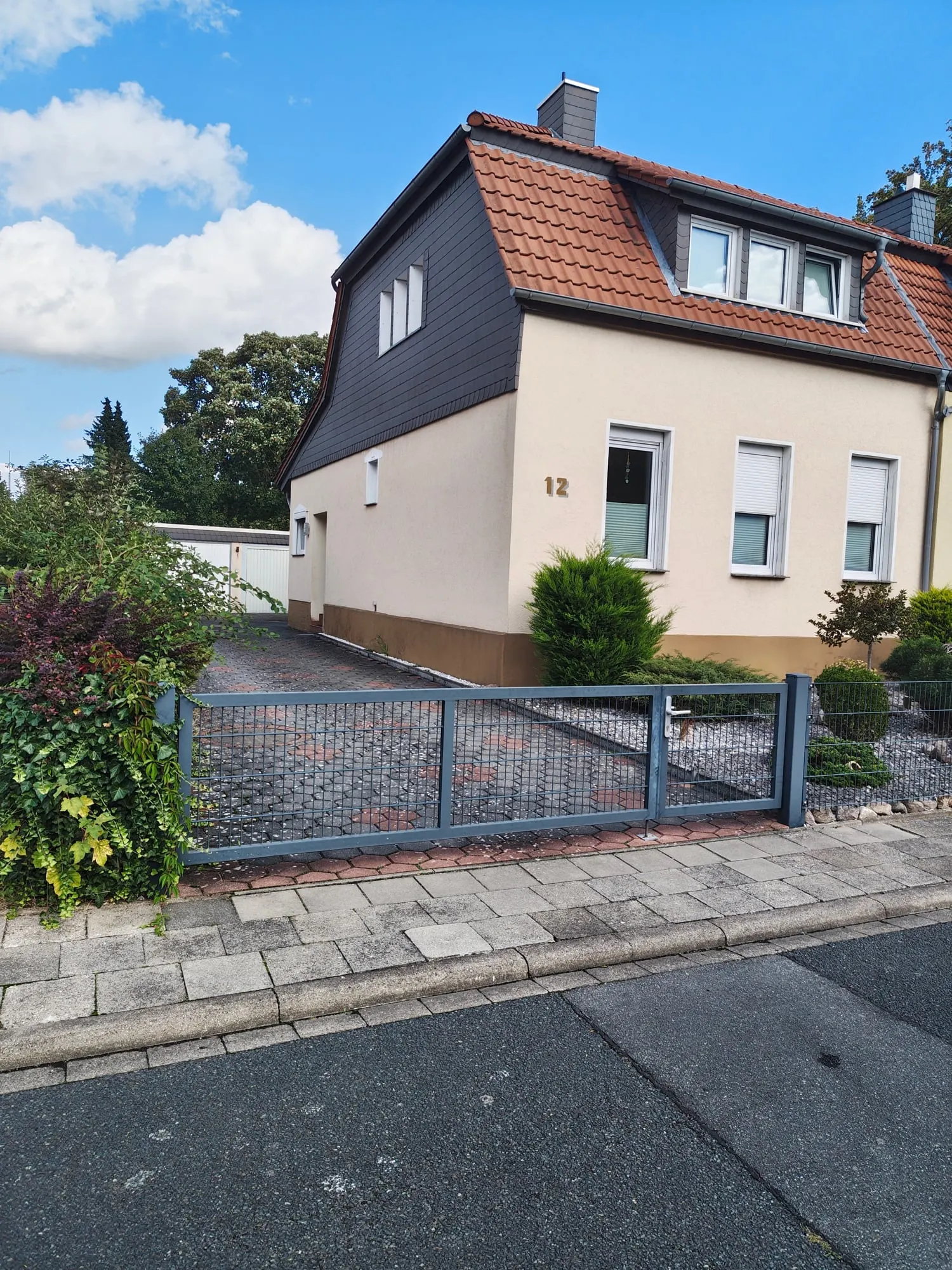 Das Haus von der Straße aus: heller Putzbau mit Gartentor, davor Hecke und Büsche.