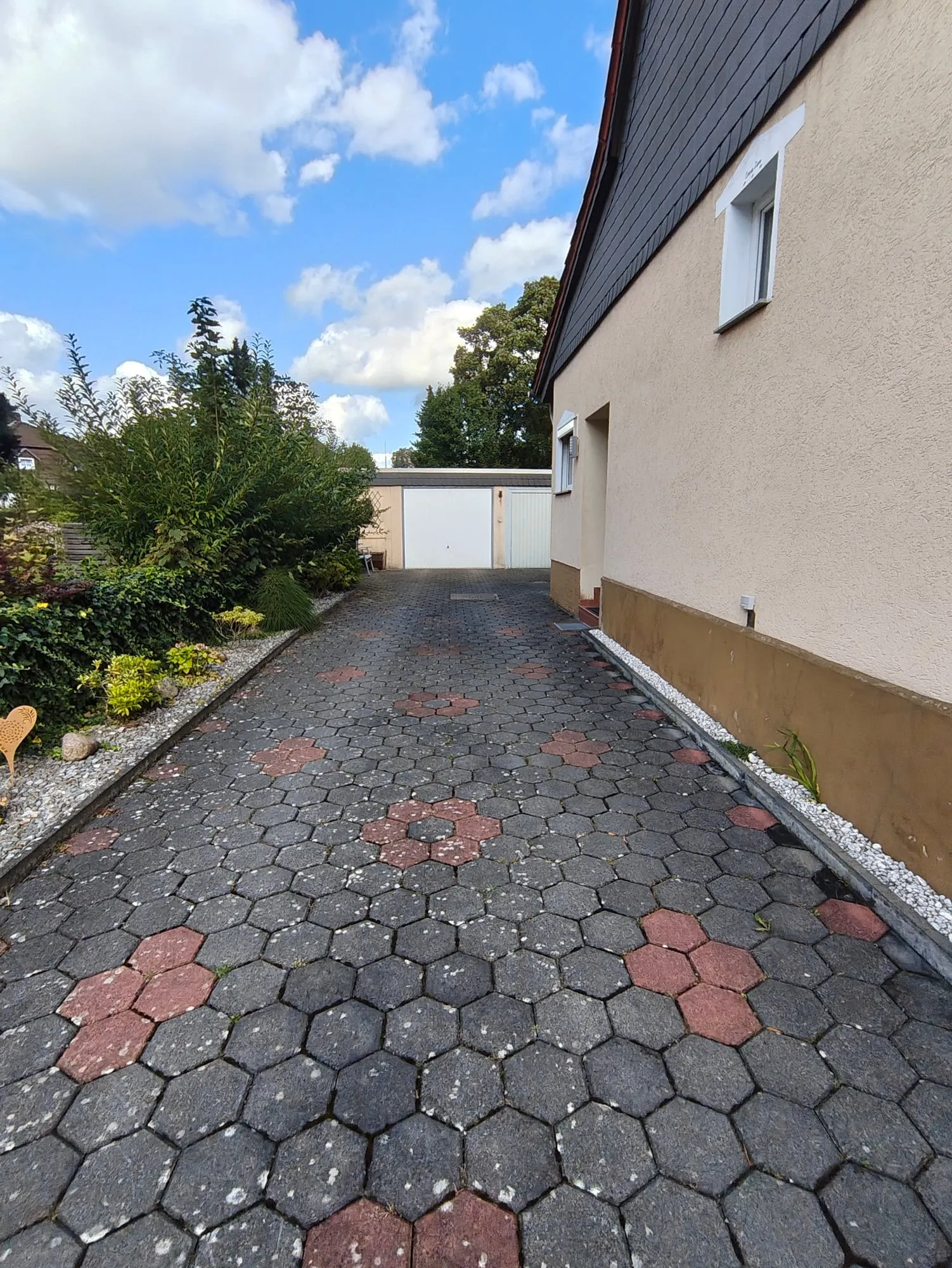 In der gepflasterten Einfahrt, links Büsche, rechts die Hauswand, vorn die Garage.