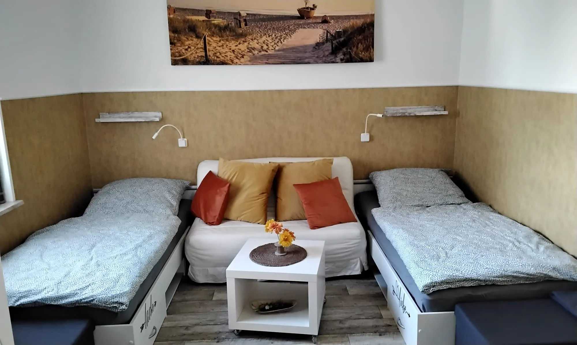 Zimmer mit zwei Einzelbetten links und rechts, dazwischen ein kleines Sofa mit orangefarbenen Kissen und ein Beistelltisch; darüber zwei Leselampen und ein Strandbild.
